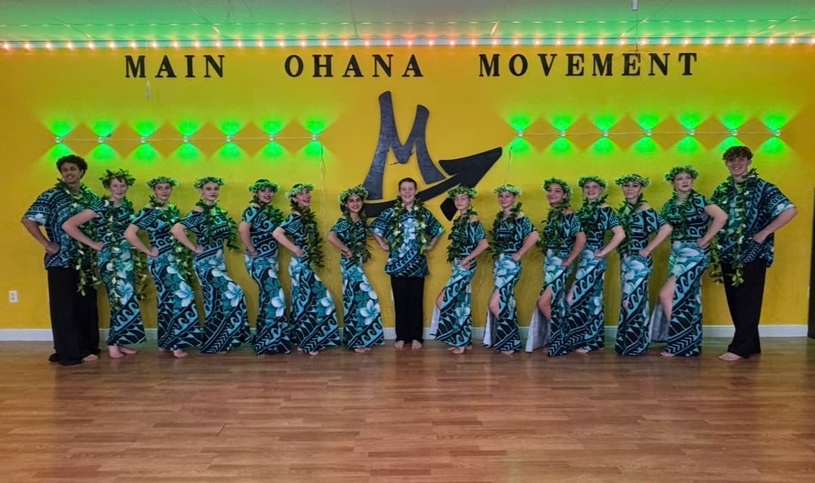 Hula dancers in matching teal print with leaf lei, lined up along the studio wall