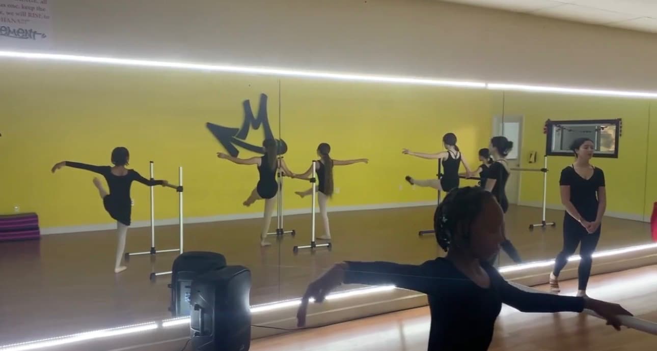 Ballet dancers in black leotards at the barre, arms extended in arabesque along the studio's mirrored wall