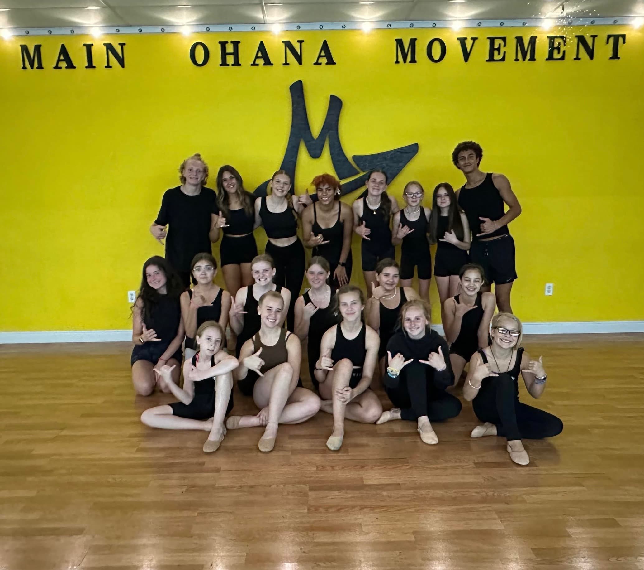 The Contemporary class in black dancewear, throwing shakas together in front of the studio's yellow logo wall